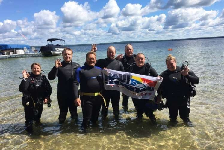 A group of scuba divers standing together in shallow lake water with a NAUI flag