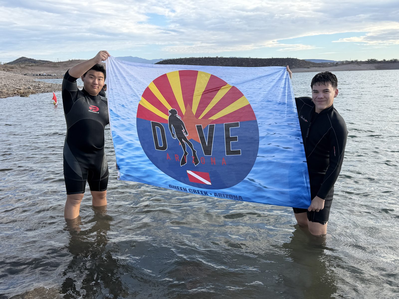 Two teen divers in wetsuits holding a Dive Arizona flag at a lake near Queen Creek, Arizona
