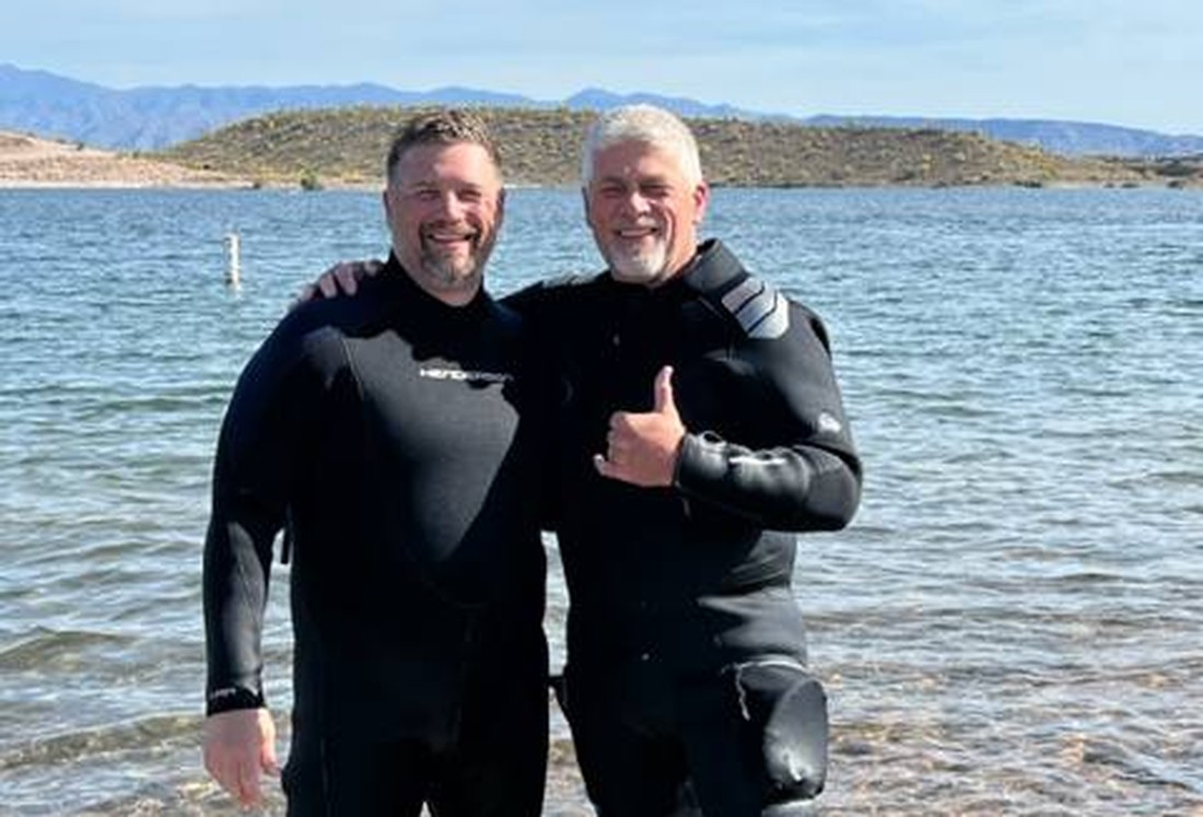 Two divers in drysuits standing together on the shore of a lake