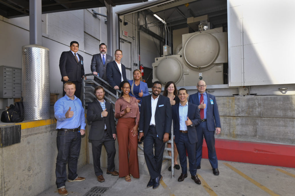 Three women and 8 men in office attire standing on an exterior staircase giving thumbs up in front of HVAC cooling machinery