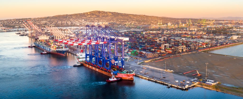 Tug boats nudge large cargo ships underneath ship-to-shore container cranes for offloading at the Port of Los Angeles at sunset. Signal Hill is visible in the distance.