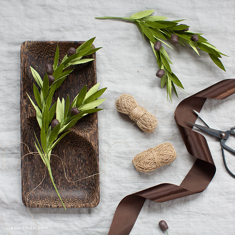 crepe paper olive branch on wooden decorative tray with brown ribbon, twine, scissors, and olive branch