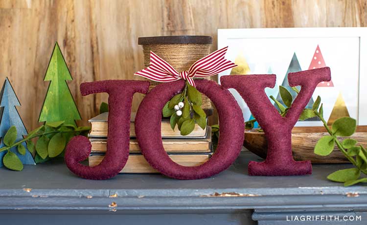 JOY felt word with crepe paper mistletoe on mantel next to books, mini trees, and framed holiday art