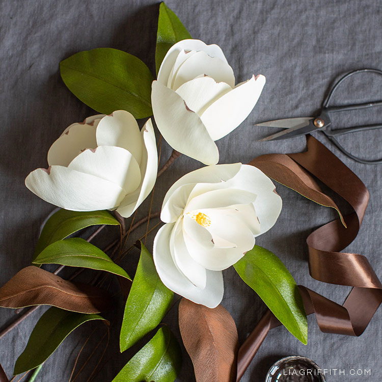 crepe paper magnolias on grey sheet next to brown ribbon and scissors