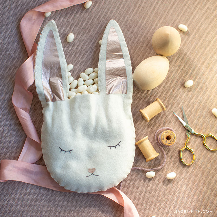 felt Easter bunny treat bag filled with candy next to pink ribbon, wooden eggs, scissors, and spools