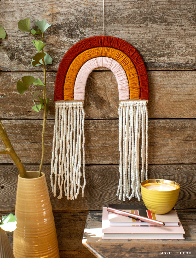 yarn-wrapped rainbow wall hanging on wood slats next to yellow vase with branch