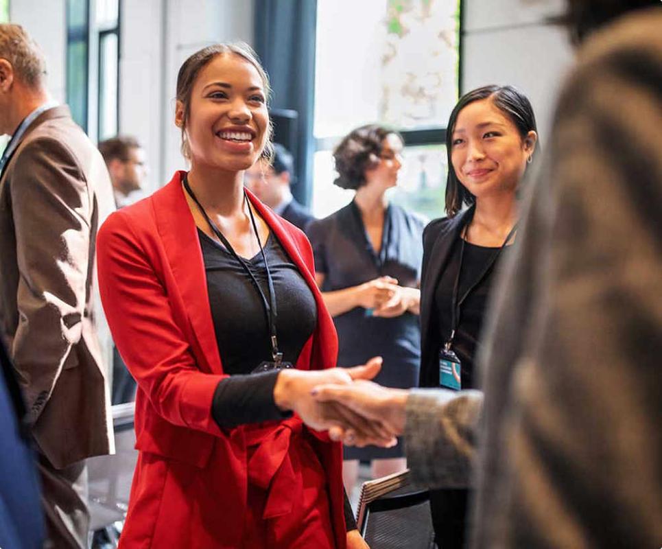 People networking at a professional event, with a woman in a red blazer smiling and shaking hands in the foreground.