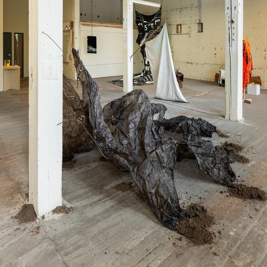 Art installation with crumpled dark material, soil, white pillars, and various textile artworks in a brick-walled room.