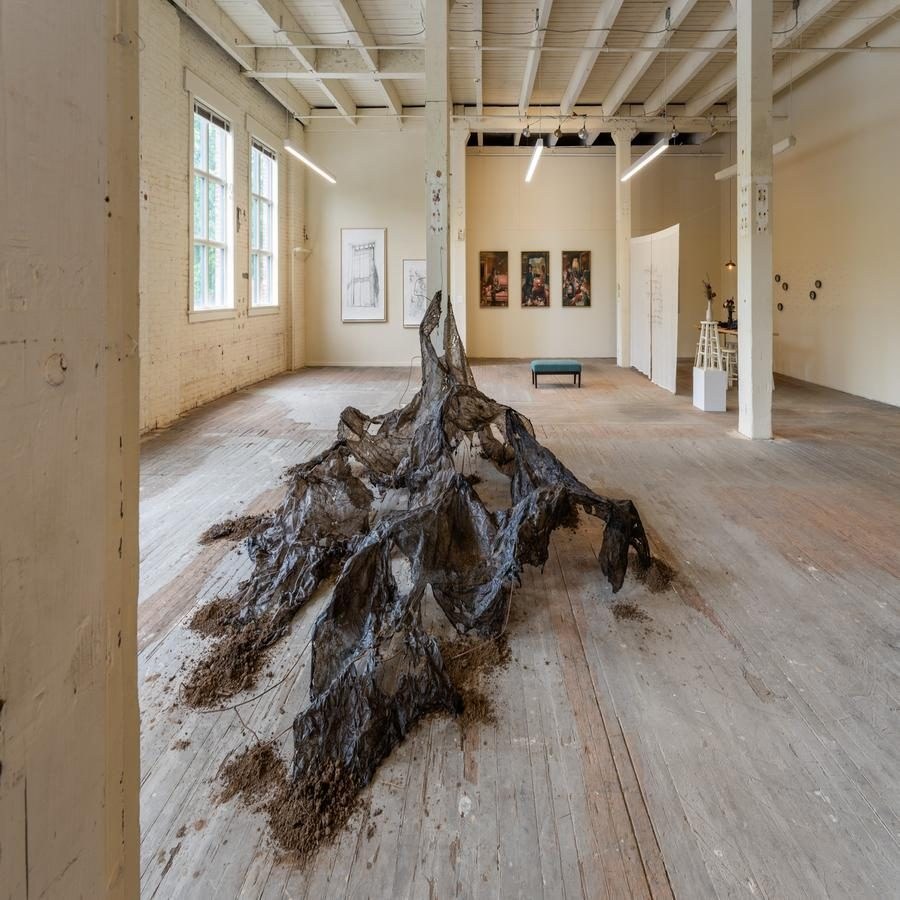 Art exhibition in a rustic room with a floor sculpture and multiple framed artworks.