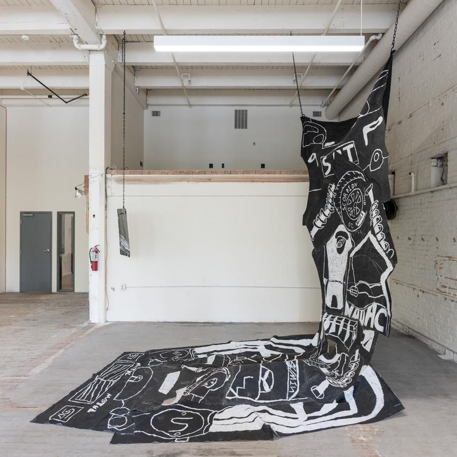 Art installation of large black fabric with white abstract designs, hanging and spreading on the floor in an industrial space.