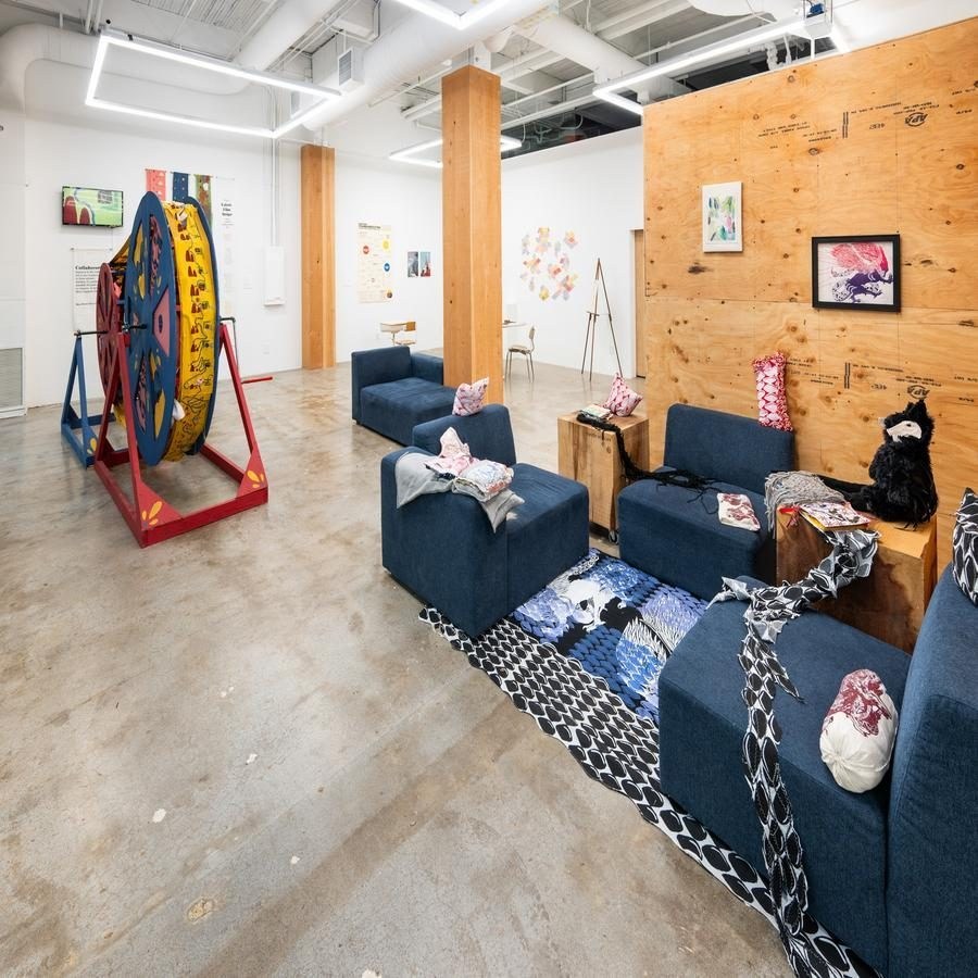 Art exhibition space with blue couches, colorful fabrics, artwork, and a large wooden wheel.
