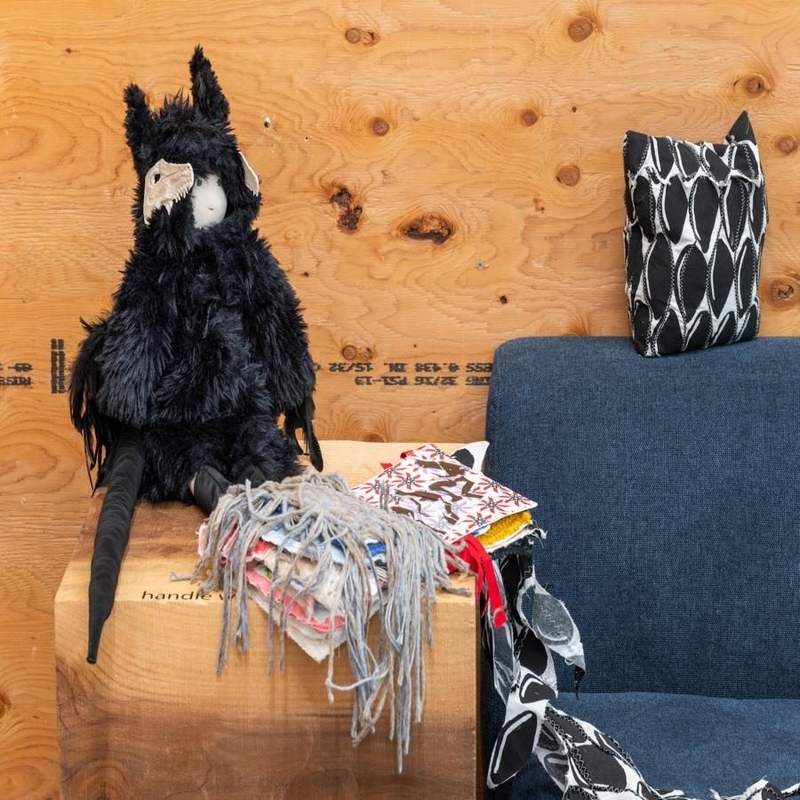 An art display with a black furry doll, colorful fabric swatches, and a patterned cushion against a wooden backdrop.