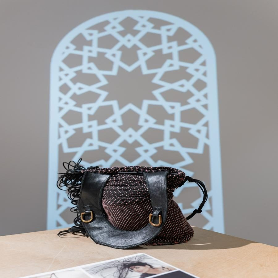 A woven handbag with black leather straps and brass rings, set in front of a blue geometric wall pattern.