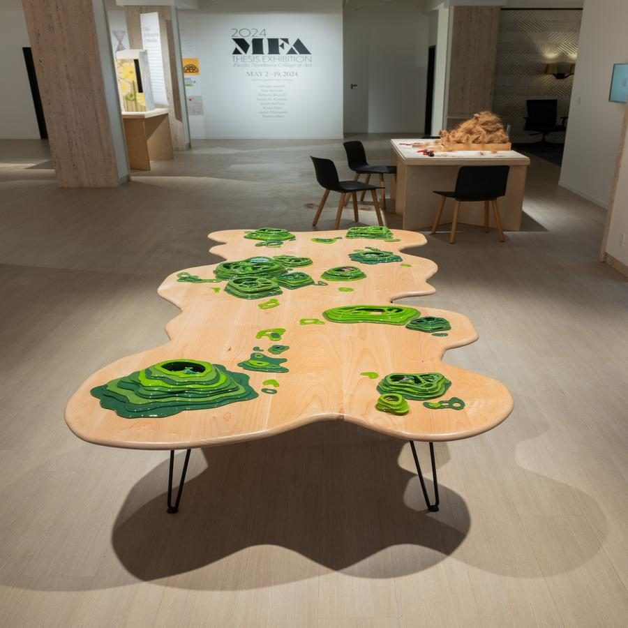 An art installation with a wood table featuring layered green topographic designs in an exhibition space.