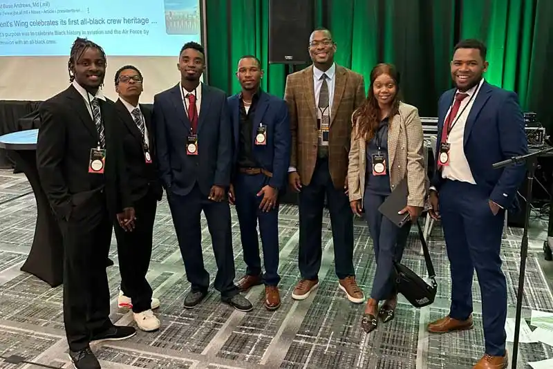 Group of seven people in formal attire wearing conference badges standing in front of green curtain.