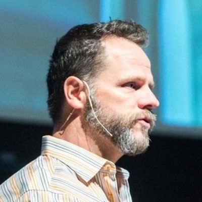 Photo, Derek Featherstone, while speaking at a conference, shown side profile with beard and over-the-ear microphone.