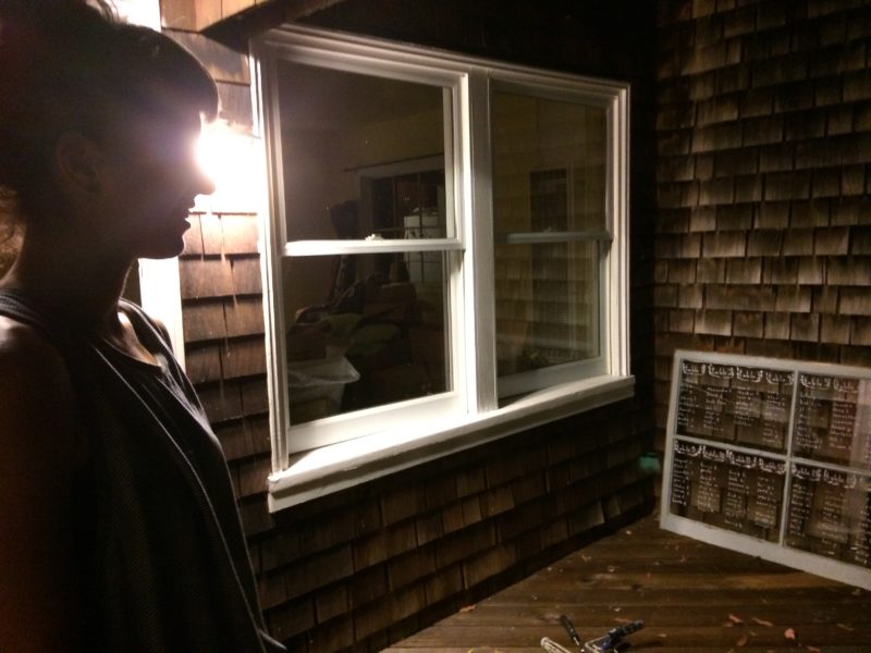 A shadowed outline of a person stands in front of the window of a weathered wooden-shingled building. Across from the person another window with white writing on it leans against the wall.