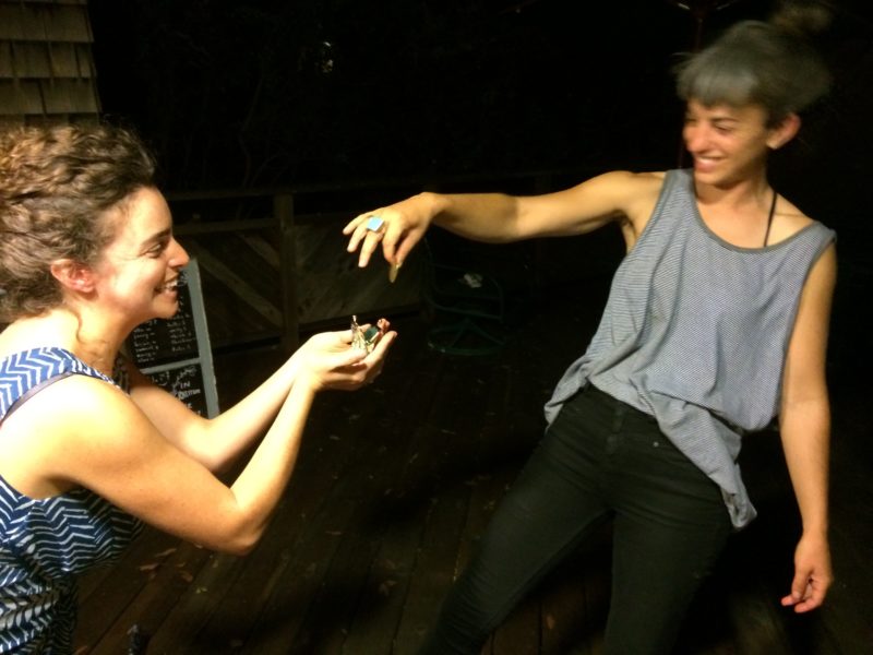 One person drops a small, unidentifiable object into the waiting hands of another person. Both people are smiling. It is dark in the background.