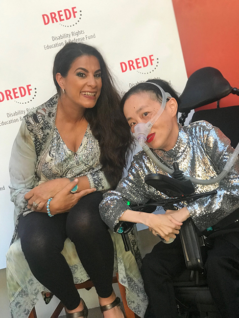 Photo of Maysoon Zayid, a Palestinian American woman with long black hair swept to one side, wearing a print top and black leggings, with Alice Wong, an Asian American woman in a wheelchair with a silver sequined top and a mask over her nose attached to a tube. Behind them is a white backdrop with the DREDF logo.