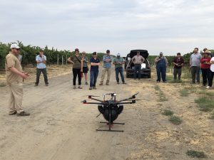 A group of people stand around a drone in the vineyard -- the drone is getting ready to lift off the ground. 