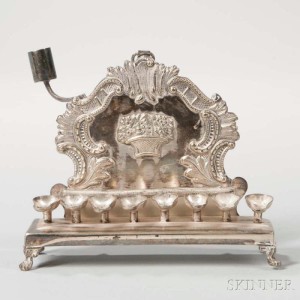 Small Polish Silverplated Hanukkah Lamp, Warsaw, late 19th century (Lot 1020). The form is elegant with mix of Rococo c-scrolls and rocaille and a Neoclassical basket of fruit, the oil pans are of minimalist design.