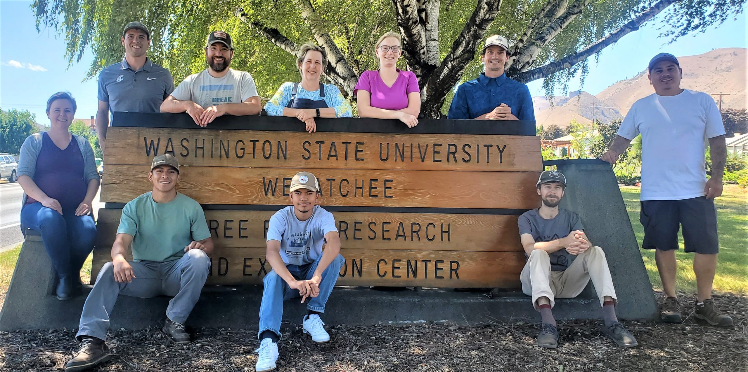A group of people from Nottingham lab are standing behind, next to and in front of a very large wooden sign with Washington State University Wenatchee Tree Fruit Research and Extension Center in black letters