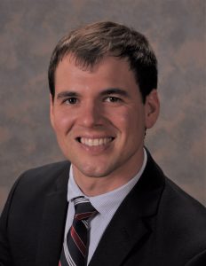 head shot of a smiling man with dark hair wearing a dark suit with a white shirt and striped tie