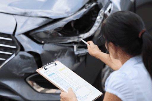 Woman inspecting front-end car damage while holding an insurance assessment form.