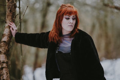 A red-haired woman dressed in a black open jacket leaning her hand against a tree, and looking away and down in disappointment.