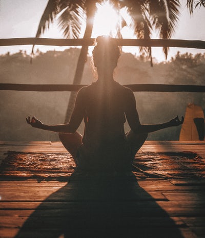 a woman whose back view we have in a meditation pose with her elbows on her knees, facing a sunrise and a palm tree.