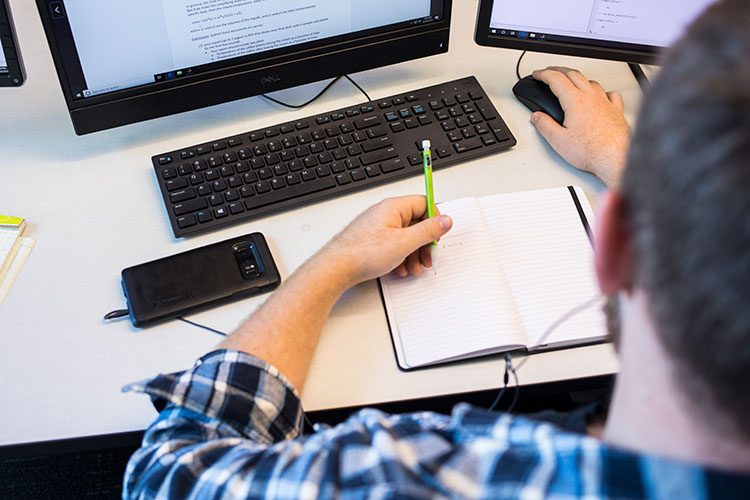 Person writing notes on paper in front of a laptop.