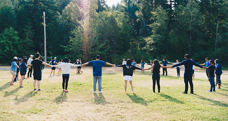 Students holding hands in a circle in a field near the woods at a Unite UW retreat