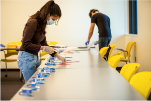 Teaching assistants prepare course materials while wearing face coverings and gloves, and staying at least 6 feet apart.