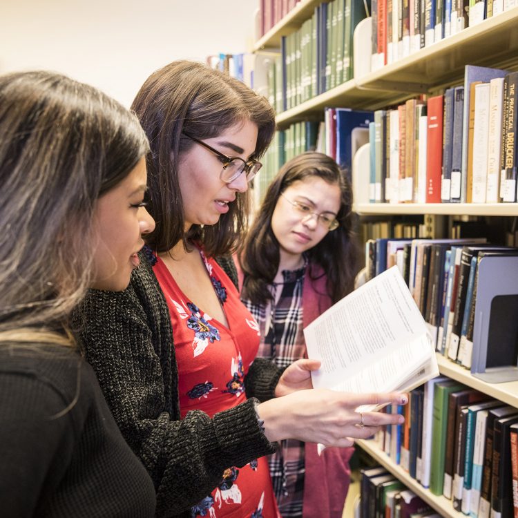 People look at an open book next to a bookcase.