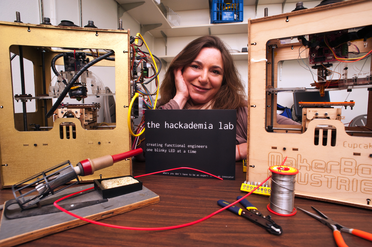 Beth Kolko with two 3-D printers built by past classes. She made the course sign herself, and came up with the class motto as she was programming the design.