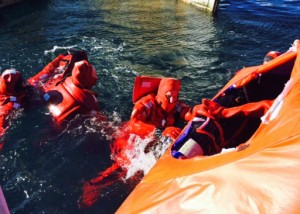 Two simmers in survival suits in water trying to climb into opening of covered life raft