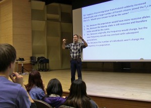 Man stands on stage in front of class, behind him is screen with questons