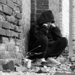 Black and white photo of boy sitting alone outside a brick building.