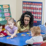 A multi-ethnic group of elementary age children are sitting at a table with their teacher and are making shapes and crafts.