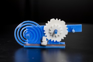 The 3-D printed gears (in white) and spring (blue spiral) toggle a switch (white box with grey surface) made of conductive plastic. The switch changes the reflective state of a 3-D printed antenna (gray strip) to convey useful data to a WiFi receiver. The shape of the gears and the speed at which they move encode the digital data.