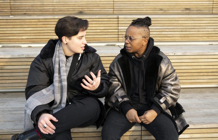 Two gender nonbinary people sit on steps outside and talk.