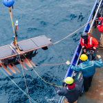 researchers on ship lowering large surfboard into water