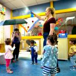 Toddlers stand in a circle, clapping. Adults stand nearby.