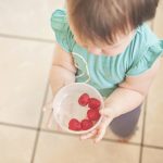 Toddler offers bowl of raspberries to camera.