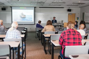 Student at a lectern with a screen behind him and audience members scattered around the room.