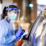 A UW Medicine worker wearing personal protective equipment stands outside a car at a drive up testing clinic