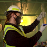 Man in hard hat and gloves in small room holding large vial with clear light brown fluid in it.