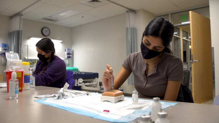 student practices inserting a syringe into a flesh-colored arm model on a table