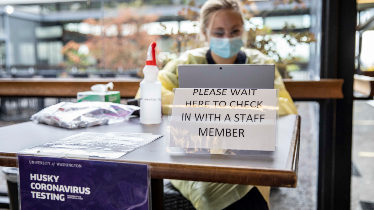 person sits behind table with sign that reads "Please wait here to check in with a staff member"