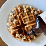 Maple syrup is being poured on a round waffle on a white plate.